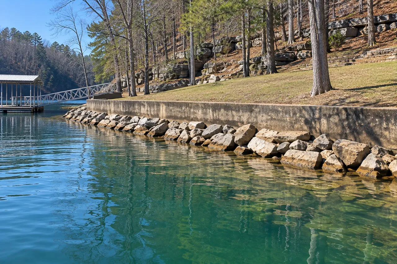 A low lake seawall with a band of riprap stone along its base, a lawn above and a dock ramp beyond
